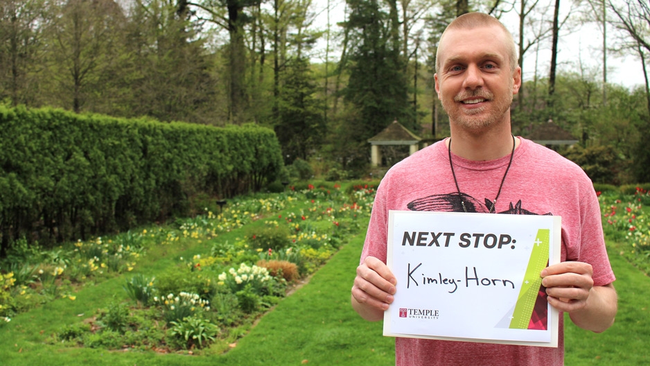 A man in a garden holding a sign that reads Next Stop: Kimley Horn.