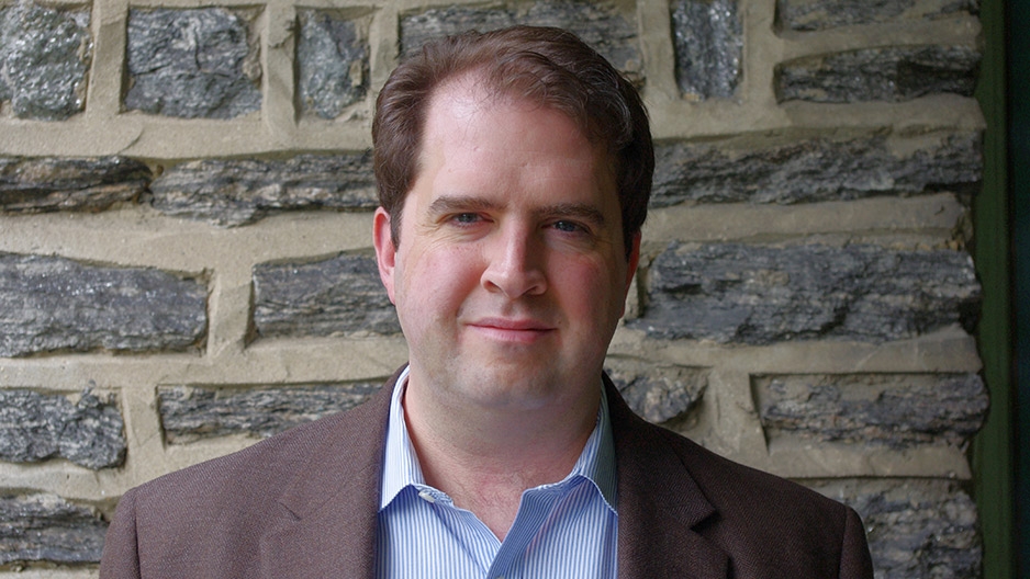 A man in a brown blazer standing in front of a stone wall.