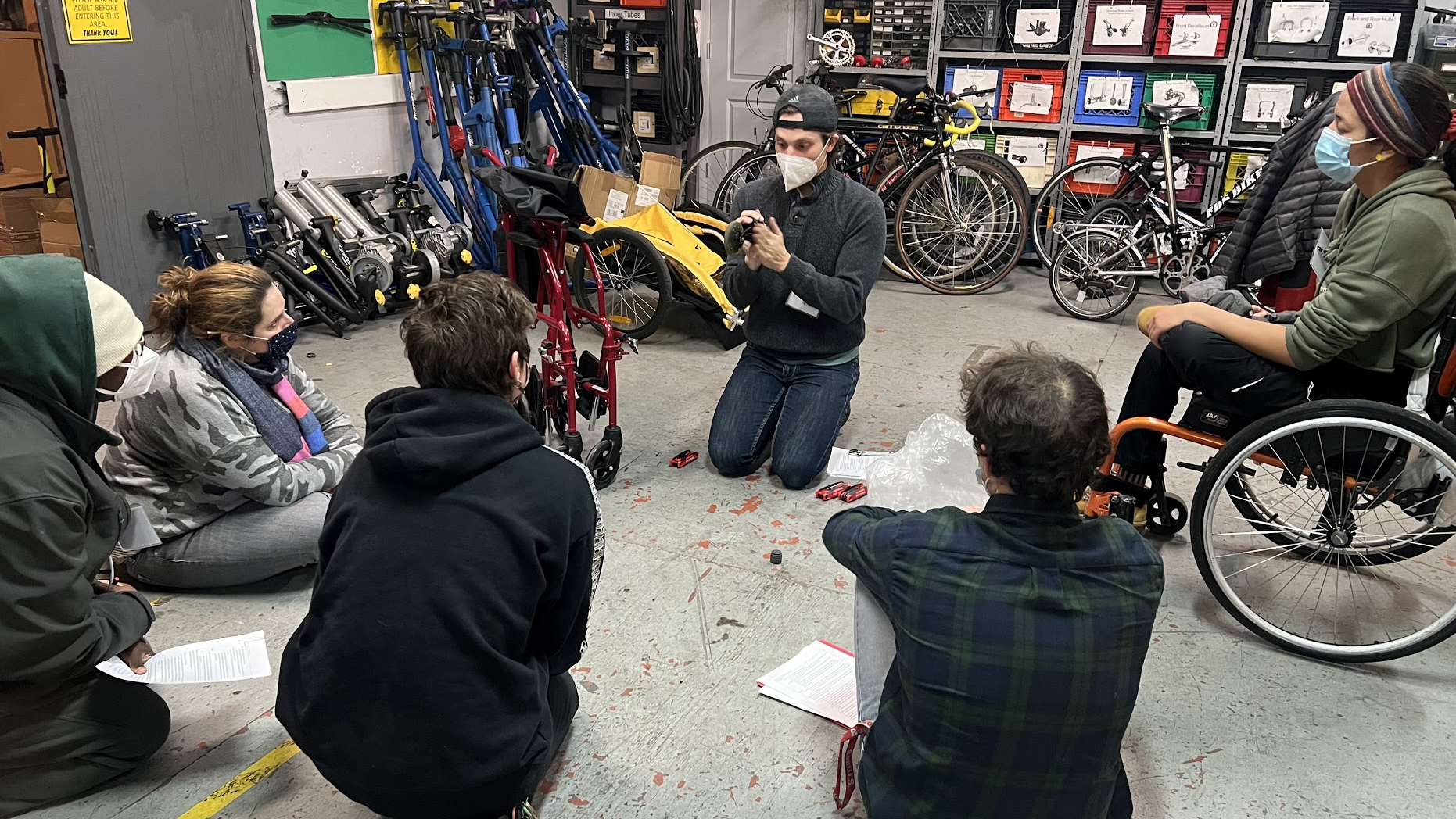 Six people at a wheelchair repair workshop. The instructor is repairing a manual wheelchair. One of the students is using a wheelchair.