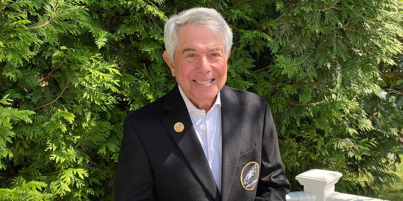 Merrill Reese holding a football and wearing his Eagles Hall of Fame jacket outside.  