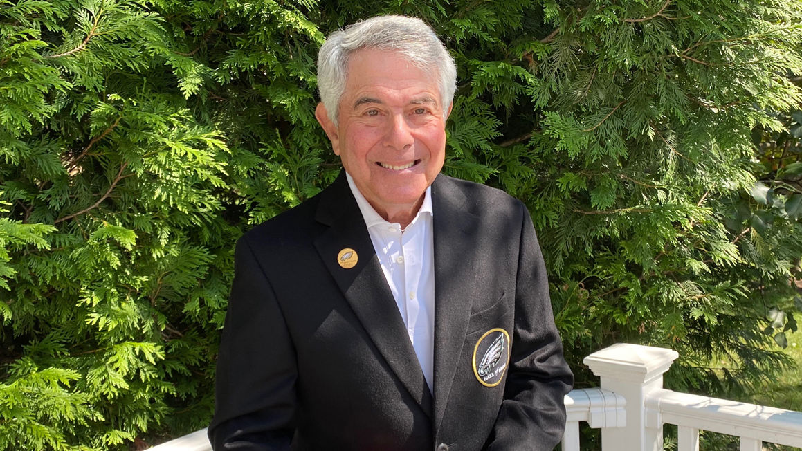 Merrill Reese holding a football and wearing his Eagles Hall of Fame jacket outside.  