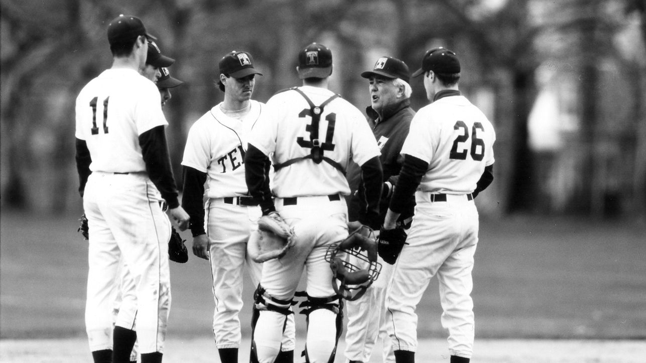 An older man with a group of student-athletes wearing caps, in black and white uniforms, standing on a raised dirt area at a Temple baseball game. 