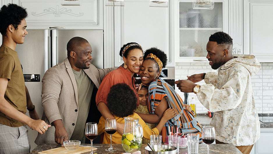 Grandparents, parents and their young children stand around the kitchen smiling and embracing.