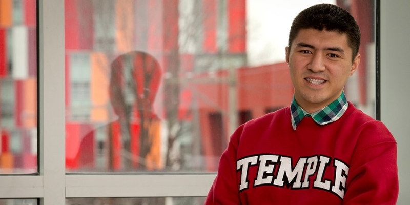 Matthew Velasquez standing in front of a window, wearing a cherry Temple sweatshirt.