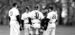 An older man with a group of student-athletes wearing caps, in black and white uniforms, standing on a raised dirt area at a Temple baseball game. 