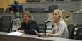 Two women sitting at a table and one is speaking into a microphone.