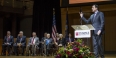 Bob Saget holding up his cell phone as he stands at a podium.  
