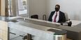 A security guard on campus wearing a mask and sitting behind a Plexiglas barrier at the entrance of a campus building. 