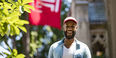 Brice Izyah smiling, wearing a red hat and standing under a Temple flag.