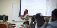 A woman in a red face covering standing before an overhead projector screen. 
