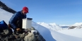 A man in a red Temple baseball cap perched on rocks with a snowy backdrop.