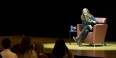 Tina Fey sitting on stage while holding a microphone and laughing. 