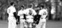 An older man with a group of student-athletes wearing caps, in black and white uniforms, standing on a raised dirt area at a Temple baseball game.