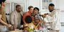 Grandparents, parents and their young children stand around the kitchen smiling and embracing.