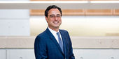 A man with dark hair and glasses wears a blue suit and smiles at a camera.