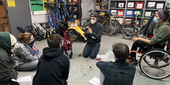 Six people at a wheelchair repair workshop. The instructor is repairing a manual wheelchair. One of the students is using a wheelchair.
