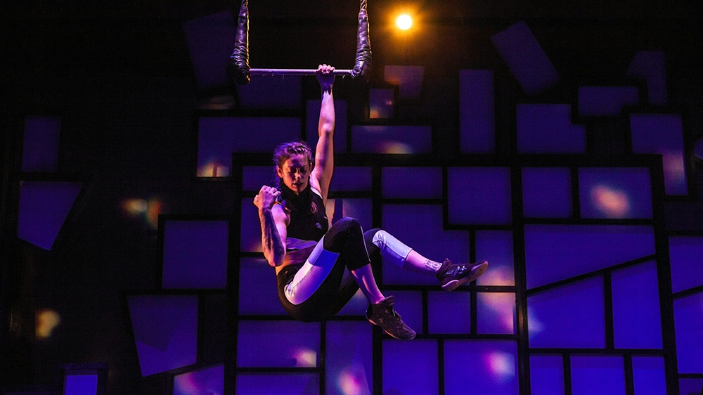 A woman hanging with one arm on a trapeze bar. 