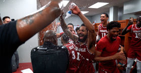 Temple basketball players wearing cherry and white jerseys, smiling brightly douse water on a coach inside the team locker room.