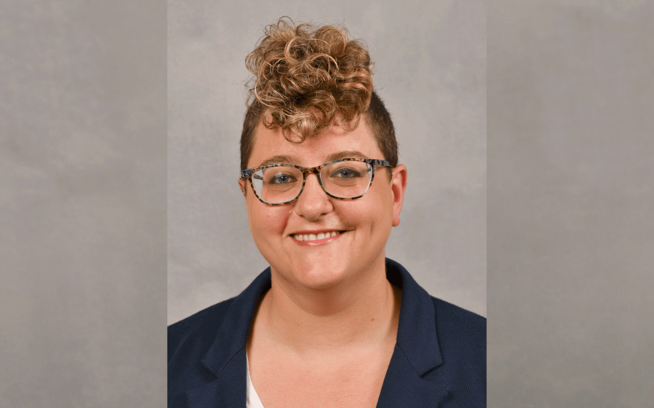 The image shows a person with short, curly hair and glasses, smiling at the camera. They are wearing a dark blazer over a white shirt. The background is a plain, neutral gray, placing the focus on the individual.