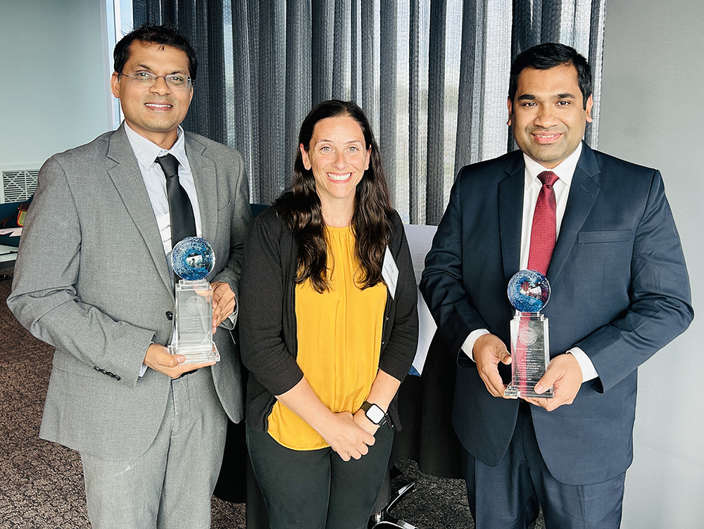 Three people. From left to right: A man in a grey suit and black tie, a woman in a black cardigan and yellow top, and a man in a navy blue suit with a red tie. They are all smiling at the camera.